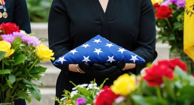 Woman holding folded American flag at military funeral service. Hands in black gloves with flowers in background. Memorial Day and Veterans Day tribute concept