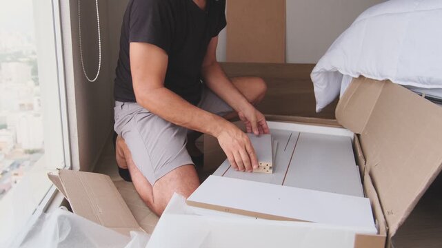 Concentrated man on his knees unboxing and examining the parts of new flat-pack furniture at home