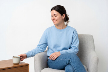 Fototapeta premium Young woman in blue sweatshirt reaching for coffee mug on table while sitting in armchair