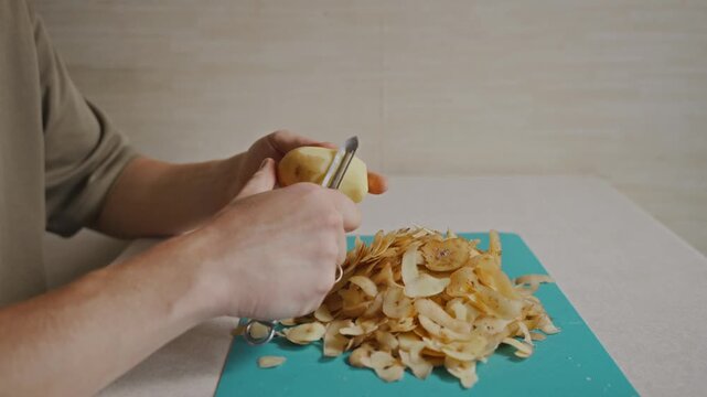 Close view of hands removing the skin from a potato with a vegetable peeler above a cutting board. Thin pieces of peel accumulate on the board during the peeling process. High quality 4k footage