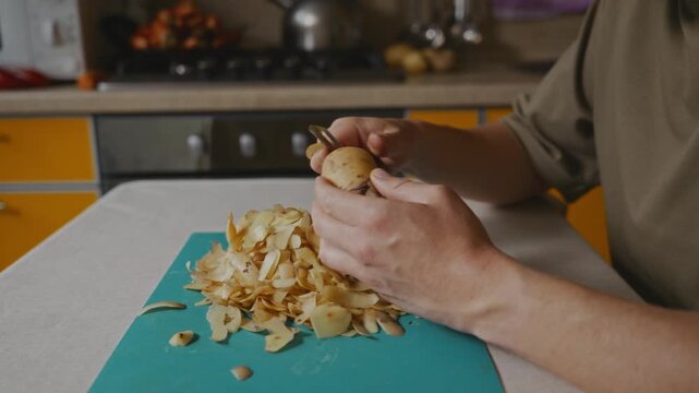 Man peeling a potato with a vegetable peeler over a cutting board. A pile of potato peels lies on the board while the hands carefully remove the skin during food preparation. High quality 4k footage