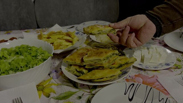 A close-up of a person's hand picking up a piece of authentic Albanian byrek with nettles and spinach from a plate, served alongside traditional side dishes like white cheese, salad, and fried potatoe