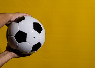 Fototapeta premium Male hands presenting a soccer ball toward the viewer with bold yellow studio background. Clear lighting highlights realistic ball surface and classic panel design, strong central