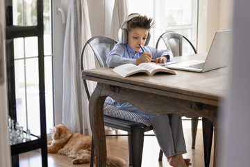 Focused child studying sitting at table with textbook and laptop, listening audio lesson through...