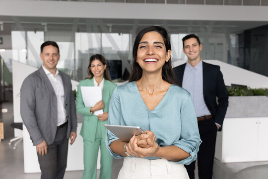Smiling young adult female team leader startup founder project coordinator standing in front of multiethnic colleagues group in office looking at camera symbolizing successful leadership teamwork