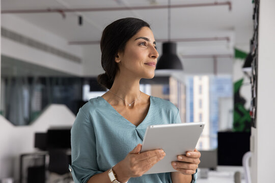 Confident Latina businesswoman planning project strategy using digital tablet in office visualizing startup success in future set goals create opportunities for personal career development and growth