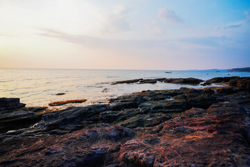 Sunset over rocky sea shore with gentle waves in tropical resort. Sun sets over ocean, casting warm colors on sea at stone rocks. Few boats can be seen on horizon as people walk along beach shore.