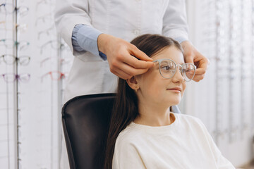 Optometrist helping young girl trying on new glasses for vision correction at optical shop