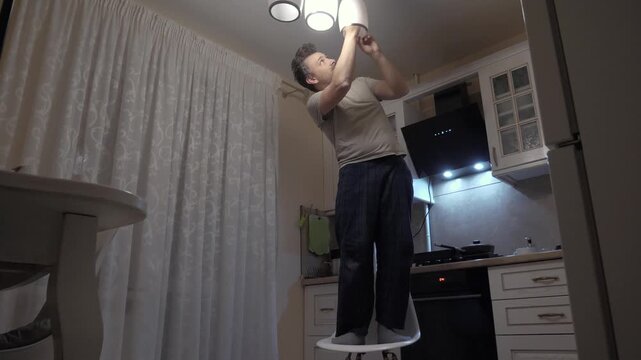Person standing on chair to change ceiling fixture. Man in casual clothes changing kitchen ceiling light fixture. Individual working to replace ceiling bulb in small apartment kitchen