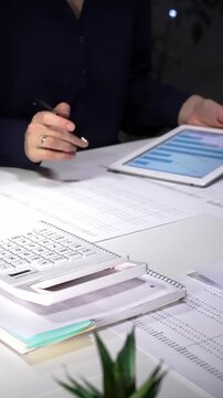 Woman working late night in office, multitasking with a tablet and writing on documents, managing heavy paperwork load. Audit and taxes in business