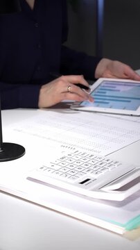 Woman working late night in office, multitasking with a tablet and writing on documents, managing heavy paperwork load. Audit and taxes in business