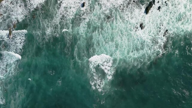 Top down aerial view of blue ocean waves crashing into rocky shore, beautiful white sea foam textures on dark water, Atlantic coast of Spain, 4K drone.