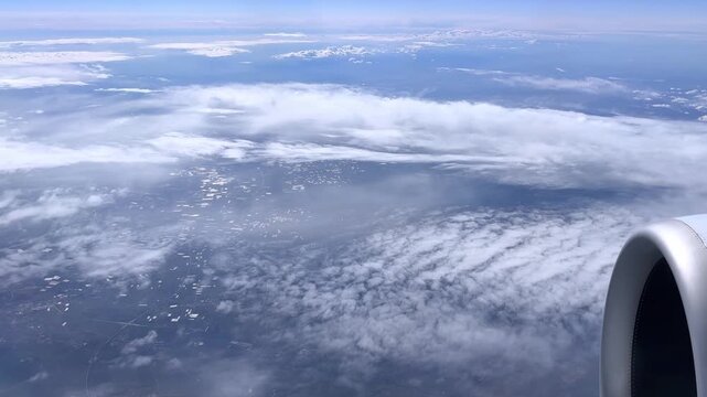 Aerial view from airplane window flying above thick layer of white clouds, Earth's landscape and ground are visible through cloud gaps under a clear blue sky, high-altitude footage of peaceful flight