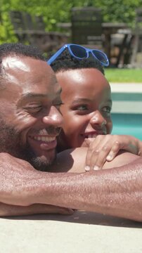 Vertical video: Clinging son urging shirtless dad leaning closer at backyard pool edge, blue shades