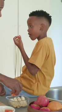 Vertical video: Eating son in mustard tee watching dad reaching for tub, making carrots at counter