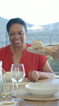 Vertical video: Hands bringing board with baguette toward senior in red blouse on patio for meal