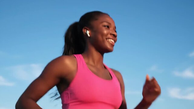Happy young adult black woman running outdoors in pink sportswear. Female athlete listening to music on wireless earbuds during a cardio workout for wellness and endurance