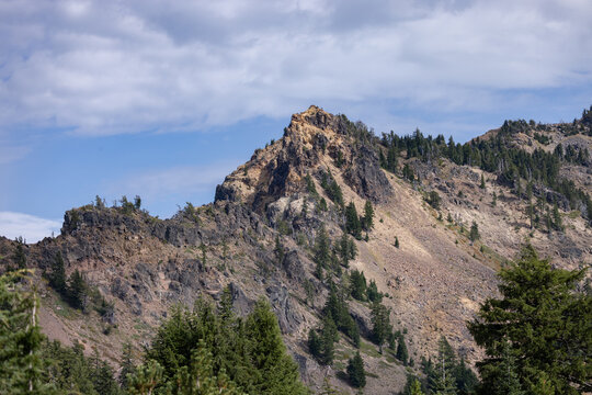 Rugged mountain peak rises sharply against blue sky. Rocky slopes host scattered pine trees and wild shrubs. Wilderness feels untouched, untamed, and serene. Crater Lake National Park in Oregon, USA.