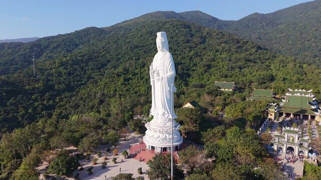 Towering White Guanyin Statue Close Aerial Showing Carving Details, Lotus Pedestal And Temple Roofs At Son Tra, Da Nang, Vietnam, Documentary Mood With Visitors As Tiny Figures And Clear