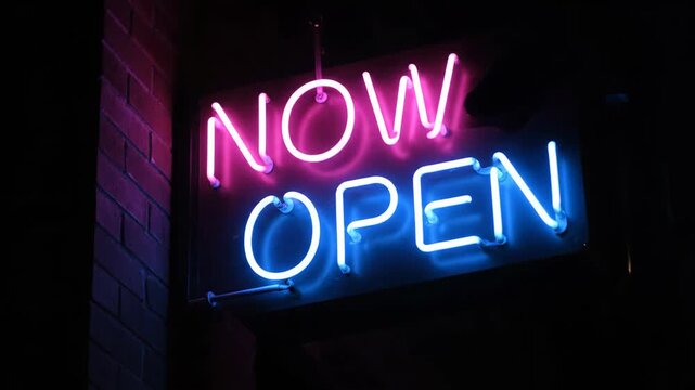 Now open neon sign glowing with vibrant pink and blue lights at night. Business advertising for a grand opening or new store welcome message. Retro urban aesthetic for commercial display