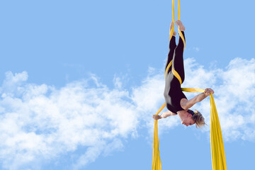 Young athletic woman hangs upside down forming a cross shape with open arms while suspended by aerial silk fabric, training outdoors to improve strength, balance, flexibility, and body control