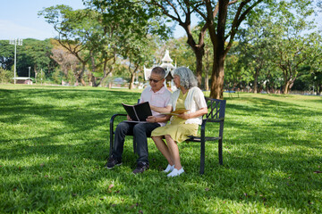 Happy asian senior couple sitting on bench in green park reading book together enjoying peaceful retirement lifestyle outdoors with smile and love