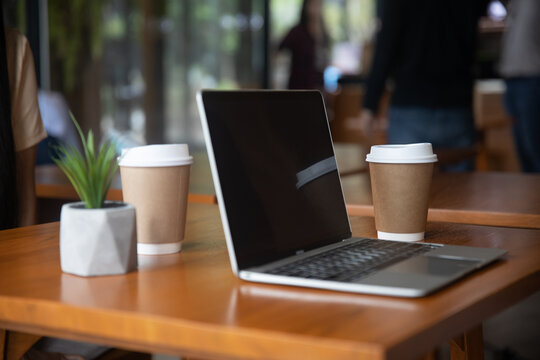 Laptop with takeaway coffee cups on wooden table in modern cafe workspace