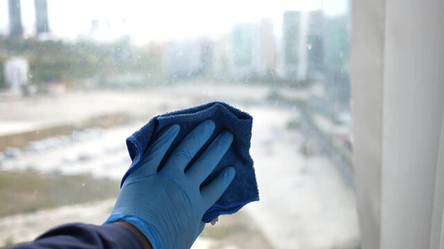 Close-up of a person wearing blue gloves cleaning a window with a microfiber cloth, showcasing the act of sanitizing and maintaining hygiene in a home or office setting with motion