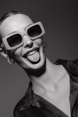 Obraz premium Young white woman with light skin and slicked hair wearing big sunglasses sticks out tongue, smiling in studio. Concept of playful mood and trendy fashion portrait.