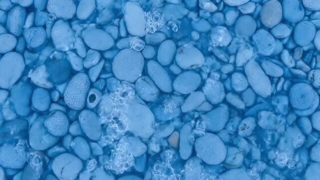 An mesmerizing overhead shot captures the gentle dance of crystal-clear water flowing over a bed of smooth, cool-toned blue and grey pebbles. The water subtly washes across the rounded stones, creatin
