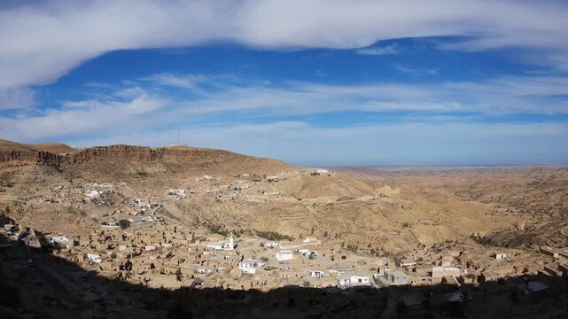Panoramic view over Toujane village in southern Tunisia, traditional houses scattered across arid hills and desert valley under dramatic clouds and wide blue sky