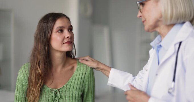 Closeup of smiling female doctor talks with older adult woman patient during medical consultation in clinic, touching her shoulder, explain procedure, share good health news, supporting and reassuring