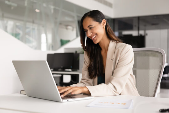 Positive businesswoman sits at desk working on laptop, lead pleasant business chat, review report with smile, feel satisfied with workflow in modern workplace. Business communication, innovative tech