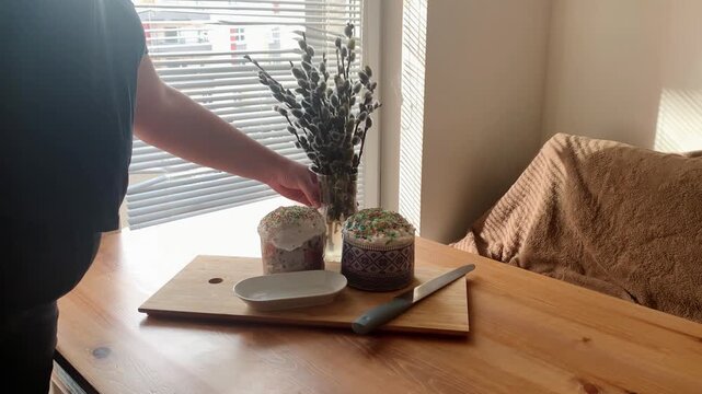 Easter table preparation with traditional Easter cakes and willow branches. Hands placing Easter cakes, willow catkins, a plate and a knife on a wooden table near a window with soft natural light. Coz