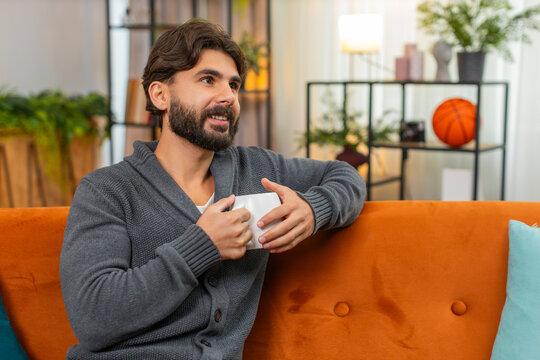 Indian man at home holds hot tea or coffee warms hands looks out window gentle smile calm. Arabian guy on sofa enjoys warm drink slow sip quiet thoughts comfort in room light cozy mood inside tonight