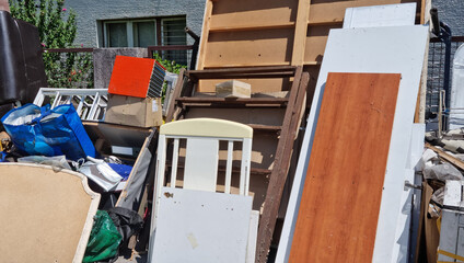 A large pile of discarded wooden boards and old household furniture sits outdoors on a sidewalk...