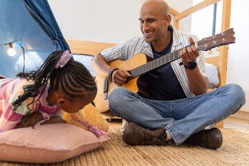 African American father and child playing guitar in bedroom, daughter holding teddy