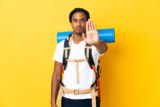 Young mountaineer man with braids with a big backpack isolated on yellow background making stop gesture