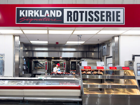 Calgary, Alberta, Canada. March 8, 2026. A Kirkland Signature rotisserie chicken station inside a Costco warehouse with a worker preparing fresh roasted poultry.