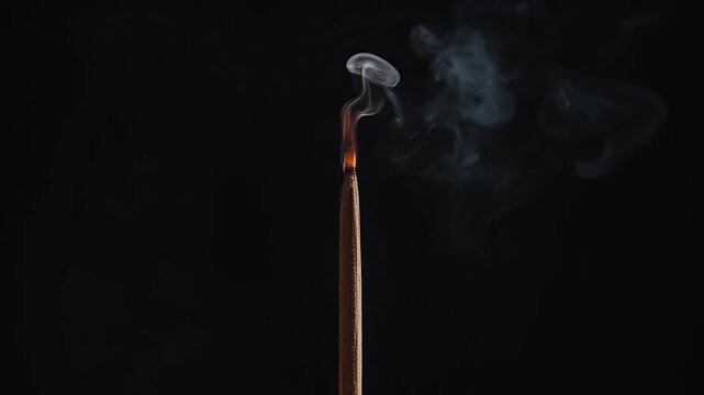 A burning incense stick emits wisps of smoke against a dark background