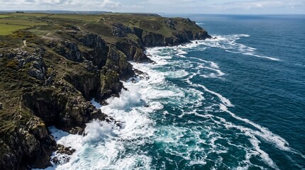 Obraz premium Dramatic aerial view of the rugged Cornish coastline with crashing waves and green cliffs under a cloudy sky.