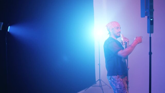 Charismatic male rapper performing a reggaeton song in a studio. The artist is singing passionately into a microphone under blue stage lights