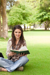 Teen female sitting cross-legged on lawn holding open green book, smiling, in jacket, backpack left
