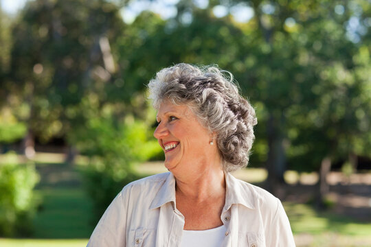 Senior woman smiling, turning right in park, wearing light button-up shirt, white top, pearl studs