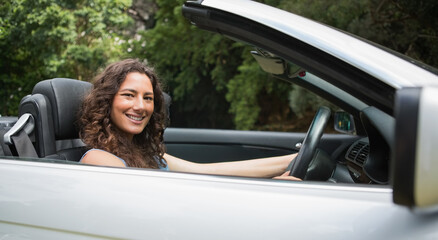 Woman wearing blue top sitting in silver convertible top down in park, reaching wheel, smiling