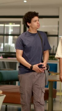 Vertical video: Student in navy tee entering left, walking lounge with phone and bag, greeting peer