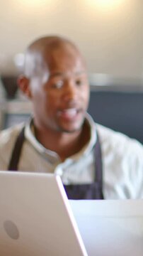 Vertical video: Answering customer, barista wearing apron using papers and laptop at cafe counter