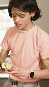 Vertical video: Turning man in pink tee pitting and scooping avocado half in kitchen preparing meal