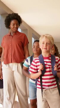 Vertical video: Walking woman in rust shirt chaperoning children along school hall with backpacks