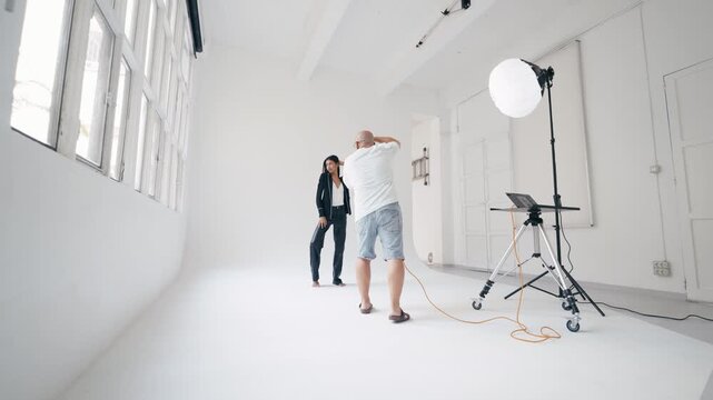 Photographer directs model posing on seamless white backdrop in bright studio, illuminated by softbox